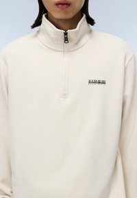 Cream zip-up sweatshirt with collar, made from soft fabric. Features a small black logo on the chest and a half-length zipper.