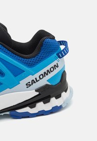 Talons de chaussures de trail de course Salomon bleu et noir avec tige en mesh, col rembourré, tirette et semelle robuste à texture multiple.