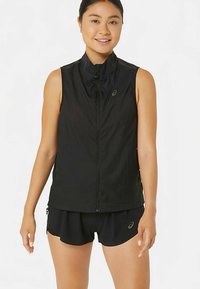 Mladá žena oblečená v bezrukavnom čiernom vestu na zips a čiernych športových šortkách, usmievajúca sa, stojaca na bielom pozadí.