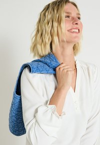 Bolso de mano acolchado azul con una textura suave y forma redondeada, llevado al hombro. Contra un fondo claro, combinado con una blusa blanca.