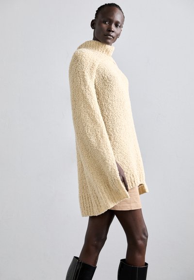 Beige knitted sweater with a high collar, textured finish, long sleeves, and side slits, paired with a light-colored skirt and black boots.