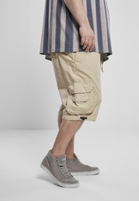 Beige cargo nadrág, oldalzsebekkel és állítható szárakkal, szürke, magasan záródó sportcipőkkel párosítva. Csíkos szürke és sötétkék póló.