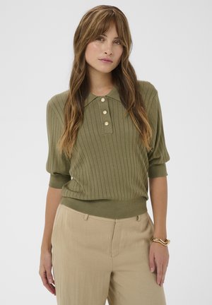 Femme aux longs cheveux bruns portant un pull côtelé à manches courtes vert olive avec des boutons et un pantalon beige sur un fond uni.