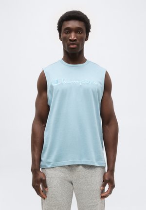 Άνδρας που φοράει ανοιχτό μπλε αμάνικο μπλουζάκι Champion και γκρι φόρμες, στέκεται με το πρόσωπο προς τα εμπρός μπροστά σε απλό λευκό φόντο.