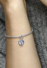 Bracciale d'argento con un design intrecciato, caratterizzato da ciondoli a forma di cuore in cristallo blu e trasparente con accenti a forma di ali.