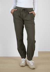 Pantalon de jogging vert olive décontracté avec poignets élastiques, taille ajustable avec cordon, poches plaquées avant, porté avec des baskets beiges et un top blanc à manches longues.