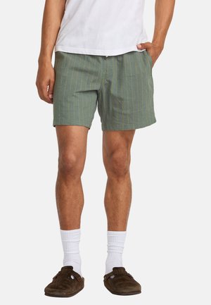 Pantalones cortos verdes con rayas amarillas, tejido ligero, corte relajado, bolsillos frontales, combinados con calcetines blancos y calzado slip-on marrón.