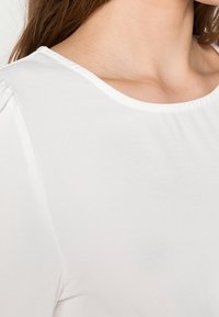 Top bianco a maniche corte con scollatura arrotondata. Tessuto liscio, spalle leggermente arricciate e materiale morbido. Nessun motivo o dettaglio visibile.