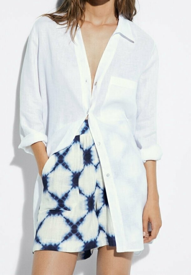 Femme portant une chemise blanche ample boutonnée sur des shorts taille haute tie-dye, les mains dans les poches, devant un fond uni.