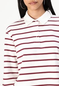 Camisa polo de manga larga, blanca con rayas horizontales burdeos, con cuello, presenta una tapeta de botones y una textura suave.