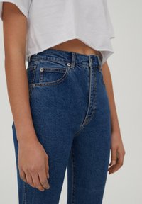 Personne portant un jean bleu taille haute et un t-shirt blanc court, debout avec les bras détendus le long du corps devant un fond uni.