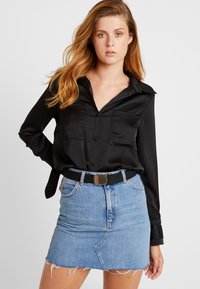 Camisa negra de satén de manga larga con cierre de botones y detalles de bolsillo, combinada con una falda corta de mezclilla con dobladillo deshilachado y cinturón negro.