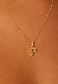 Gouden halsketting met een gestructureerd hangertje in de vorm van de letter "P". De ketting is fijn en delicaat, liggend op een warme huidtint achtergrond.