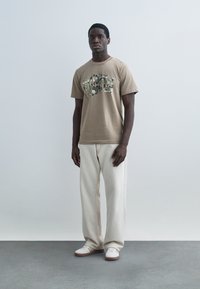 Jeune homme debout portant un t-shirt beige à motif, un pantalon large crème clair et des baskets blanches, sur un fond gris uni.