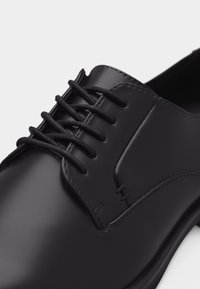 Μαύρο στιλβωμένο δερμάτινο παπούτσι φορέματος με κλειστό κορδόνι και στρογγυλεμένη μύτη, εμφανιζόμενο σε κοντινή λήψη με λευκό φόντο.