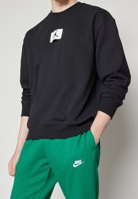 Sudadera negra con puños acanalados, que presenta un parche de logo blanco en el pecho. Combinada con pantalones deportivos verdes, que muestran un logo blanco de Nike.