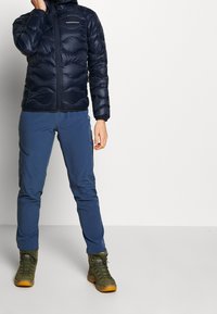 Veste rembourrée marine avec fermeture éclair sur le devant, finition texturée ; associée à un pantalon bleu clair et des bottes de randonnée vertes avec des semelles marron.