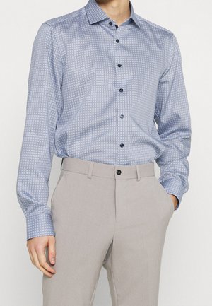 Homme portant une chemise à manches longues bleu clair à motifs, rentrée dans un pantalon beige, une main dans la poche et l'autre détendue sur le côté.