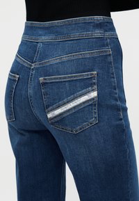 Σκούρο μπλε τζιν με πέντε τσέπες, ραφές σε αντίθεση και ετικέτα με επωνυμία στην πίσω τσέπη. Λείο υλικό.