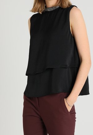 Ærmeløs sort lagdelt top med perlebesat krave båret med burgunderrøde bukser, hånd i lommen, på lysegrå baggrund.