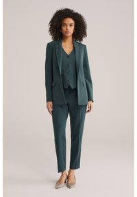 Vrouw met krullend haar, gekleed in een donkergroen op maat gemaakt pak met een gilet en beige pumps met puntige neus, staand tegen een effen achtergrond.