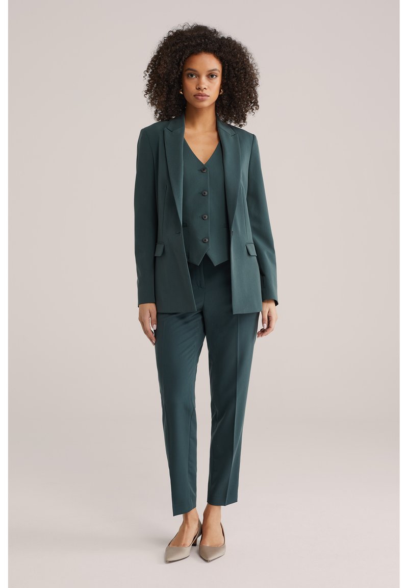 Vrouw met krullend haar, gekleed in een donkergroen op maat gemaakt pak met een gilet en beige pumps met puntige neus, staand tegen een effen achtergrond.