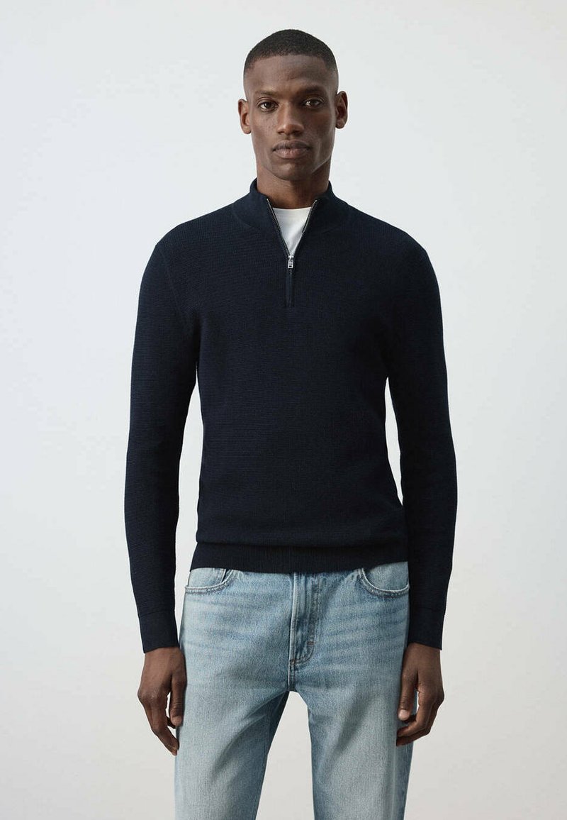 Homme portant un pull noir à demi-fermeture éclair sur une chemise blanche et un jean bleu clair, debout devant un fond clair uni.