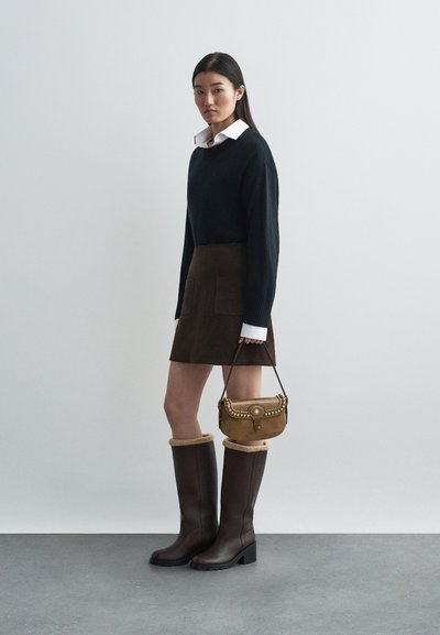 Black knit sweater, brown suede skirt with front pockets, brown knee-high boots with fur trim, and a small tan handbag with studs.