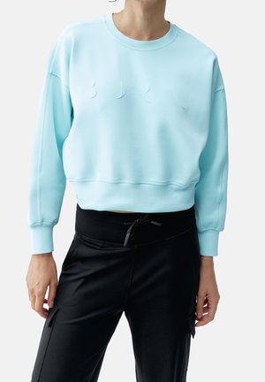 Personne portant un sweat-shirt court bleu clair avec des lettres embossées discrètes et un pantalon noir à cordon avec poches latérales.