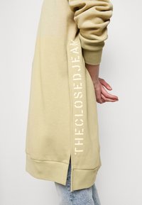 Długa beżowa bluza z bocznymi rozcięciami, z białym napisem "THE CLOSED JEAN" wzdłuż jednej strony, wykonana z miękkiego materiału.