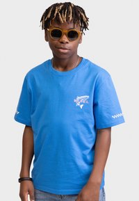 Camiseta de algodón azul con diseño de pez y texto "REBEL" en rosa, con acentos a cuadros blancos en las mangas. Usada con gafas de sol amarillas.