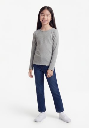 Chica sonriente de pie, con una camiseta gris de manga larga, jeans azules y zapatillas blancas, sobre un fondo blanco liso.