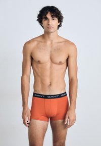 Bóxer naranja con una cintura elástica negra que presenta el logo "GANT" repetido. Tejido suave y diseño ajustado.
