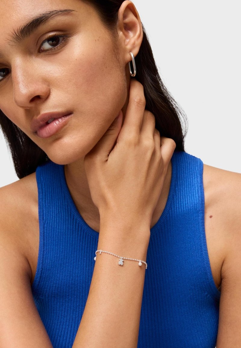 Bracciale in argento con piccoli ciondoli decorativi, indossato su un polso. Modella con un top blue a costine senza maniche, che mostra un orecchino a cerchio.