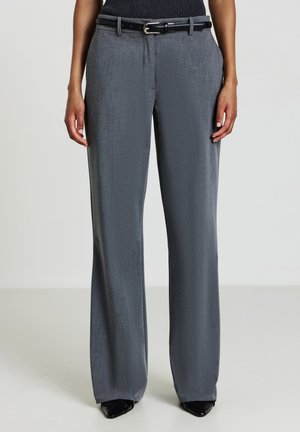 Femme portant un pantalon gris taille haute à jambes larges avec une ceinture noire et des chaussures pointues noires sur un fond uni.