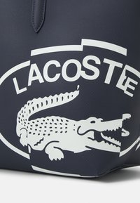 Sac cabas bleu marine en matériau synthétique texturé, doté d'un grand logo Lacoste blanc et d'un alligator en graphique bien visible.