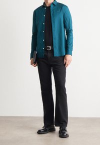 Tealblauw overhemd met lange mouwen en knopen, zwarte T-shirt eronder, zwarte jeans, zwarte schoenen, een zwarte clutch vasthoudend. Soepele stof, getailleerde pasvorm.