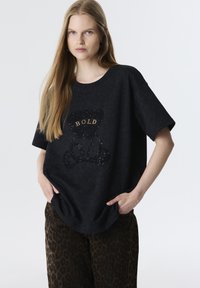 Zwart oversized t-shirt met een textuurontwerp van een beer en het woord "BOLD" in goud. Gecombineerd met bruine luipaardprint broeken.