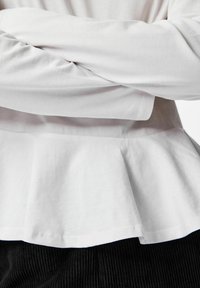 Hvit langermet topp med peplum-design, med glatt bomullstekstur og en nederkant som flerrer lett ut ved midjen.