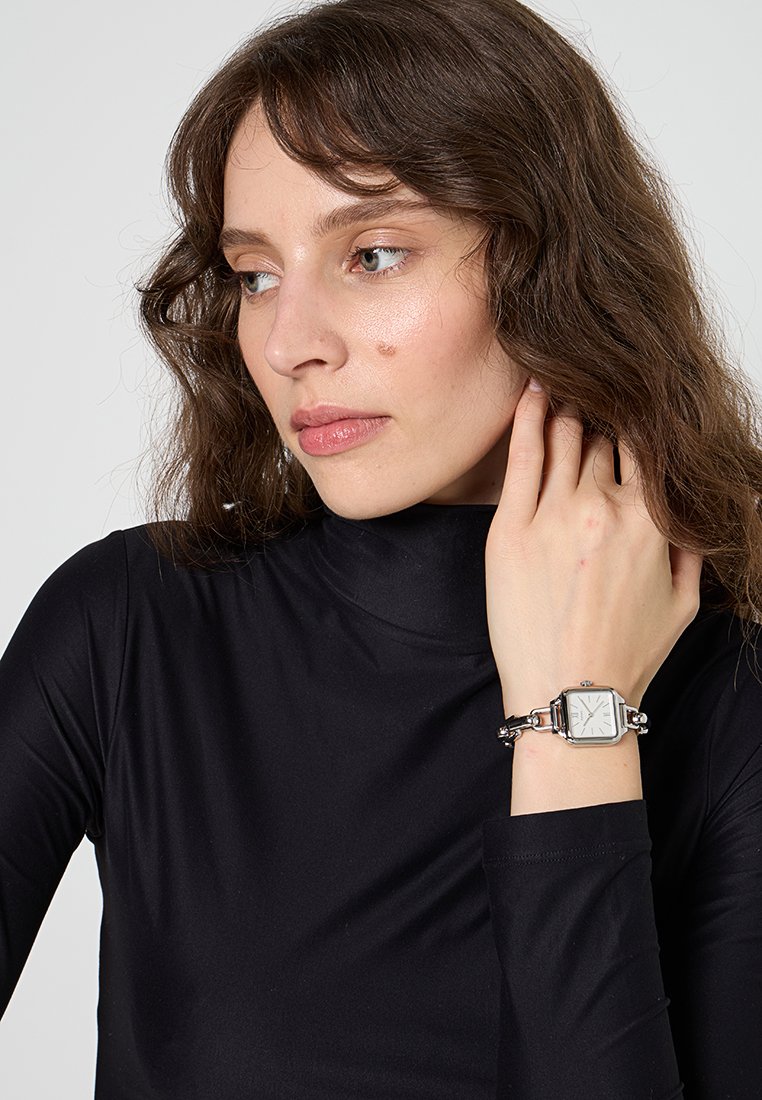 Orologio in metallo argentato con quadrante quadrato, faccia del quadrante bianca e cinturino a maglie. Indossato su un polso, abbinato a un top nero a maniche lunghe.