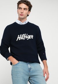 Granatowy sweter z dzianiny z białym logo "Hilfiger", okrągły dekolt i prążkowane mankiety, w zestawie z jasnoniebieskimi dżinsami. Teksturowana tkanina, styl casual.