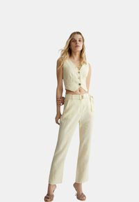 Top beige senza maniche con scollo a V e chiusura a bottoni abbinato a pantaloni beigi a vita alta con gamba dritta, dotati di cintura e tasca.