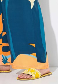Blå och orange mönstrade vida byxor i kombination med gula slidesandaler som har vit text och strukturerade detaljer på sulan.