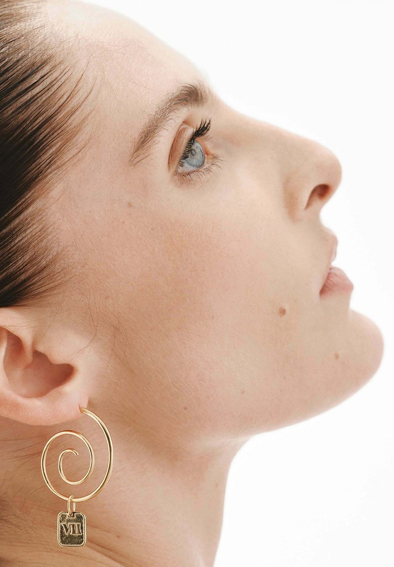 Perfil de una mujer con ojos azules que lleva un pendiente dorado en espiral con un colgante rectangular marcado con "VII" en su oreja izquierda.