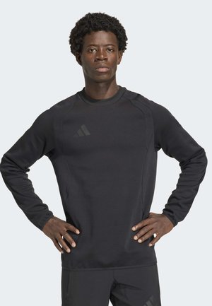 Jonge zwarte man staat met zijn handen op zijn heupen, draag een zwart, lang mouw atletisch shirt en zwarte broek tegen een eenvoudige achtergrond.