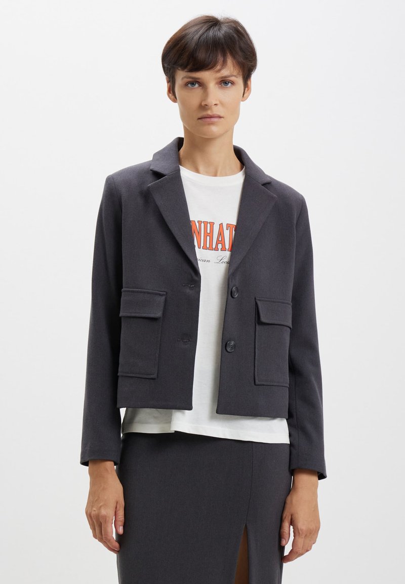 Veste grise foncée raccourcie avec revers crantés, deux poches avant et fermeture à boutons, portée sur un t-shirt graphique blanc. Texture lisse.