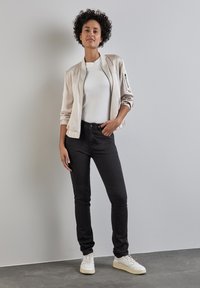 Ljusbeige bomberjacka över en vit t-shirt, tillsammans med svarta jeans och vita sneakers. Släta texturer, åtsittande siluett och minimala detaljer.