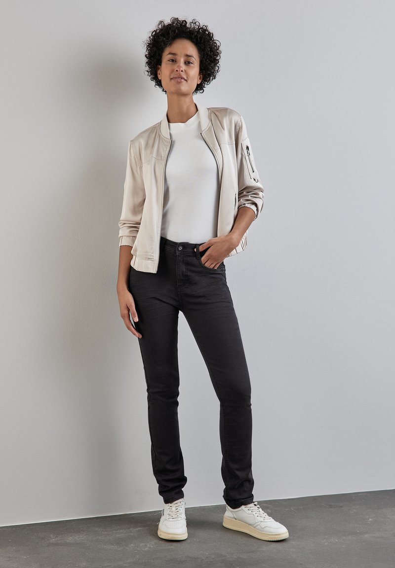 Ljusbeige bomberjacka över en vit t-shirt, tillsammans med svarta jeans och vita sneakers. Släta texturer, åtsittande siluett och minimala detaljer.