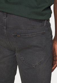 Tmavě šedé džíny z denimu s výraznou zadní kapsou a žlutým logem "Lee". Textilie vypadá hladce s kontrastním prošíváním.