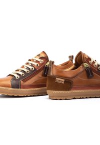 Las zapatillas de cuero marrón cuentan con un diseño texturizado, acentos oscuros contrastantes, ojales de metal y un detalle de cremallera lateral. Las suelas de goma completan el look.